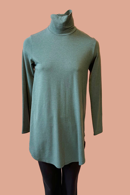 The Lauren Top by Advika in PIne, with a turtleneck, swing shape, long sleeves, and a rounded hi-low hem, is show on a mannequin in front of a coral background. 