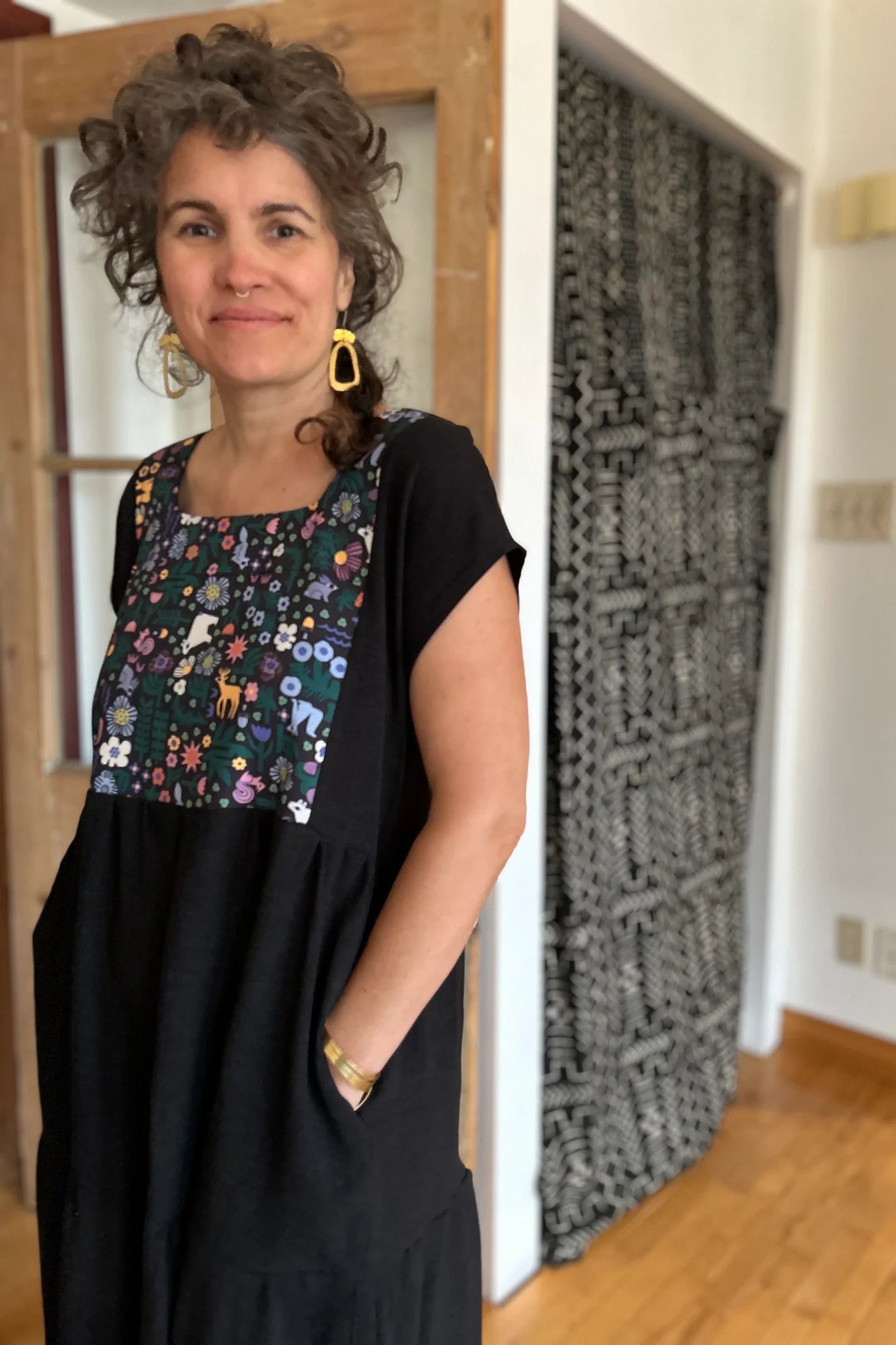 Woman wearing the Lavendula Dress by Kazak in Farmland, a short sleeved square neck midi dress with three ruffles. She is standing in a room with wooden floors and a door.