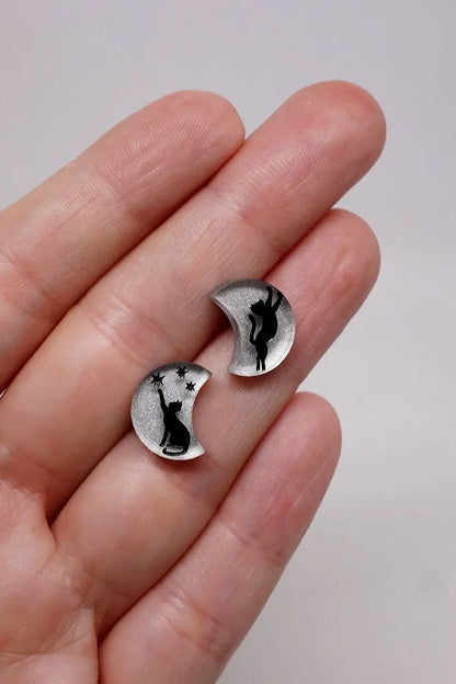 Silver moon shaped earrings with a black cat design held between fingers against a plain background/