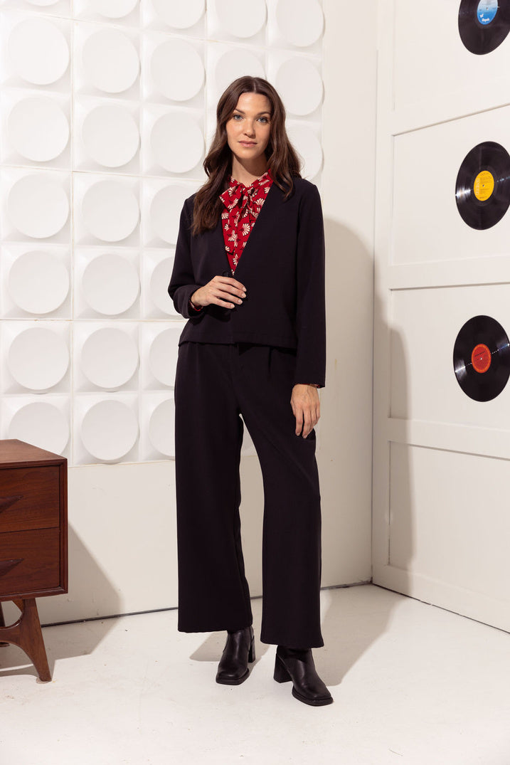 A woman wearing the Bowie Jacket by Cherry Bobin in Black featuring a V-neck, a single button closure, long sleeves and a short and slightly boxy shape. She is wearing it with a red blouse and matching pants and standing in front of a white wall with vinyl records hanging on it. 