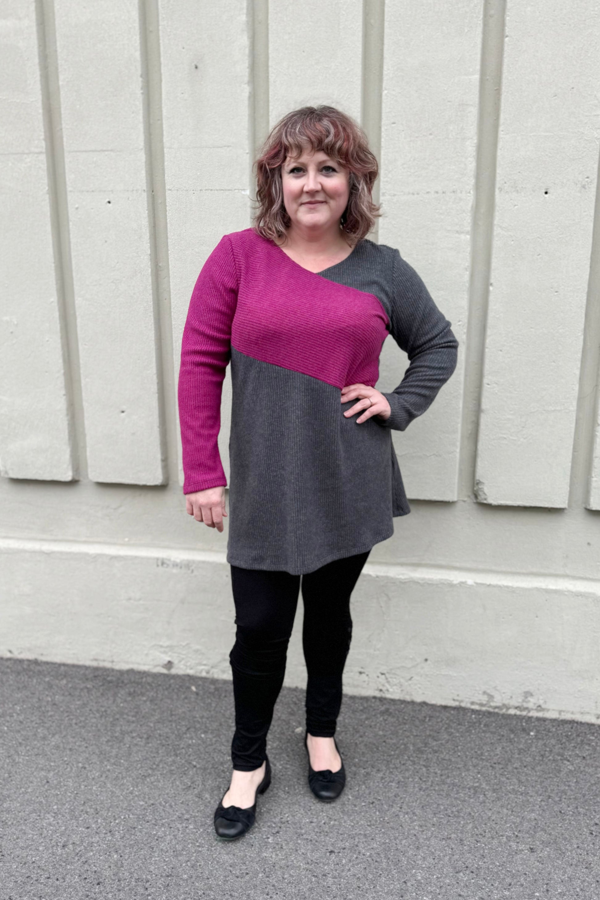 A woman wearing the Exis Tunic by Luc Fontaine in Pink/Grey, featuring asymmetrical colour blocking, long sleeves, and an A-line shape, with black leggings. She is standing in front of a white wall. 