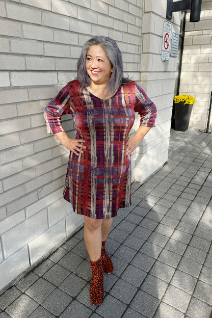 A woman wearing the Charlize Dress by Luc Fontaine in Multicolour Print in with a plaid effect in jewel tones with a touch of sparkle, featuring  a V-neck, 3/4 sleeves, and A-line shape that hits above the knee. She is standing outside in front of a brick wall. 
