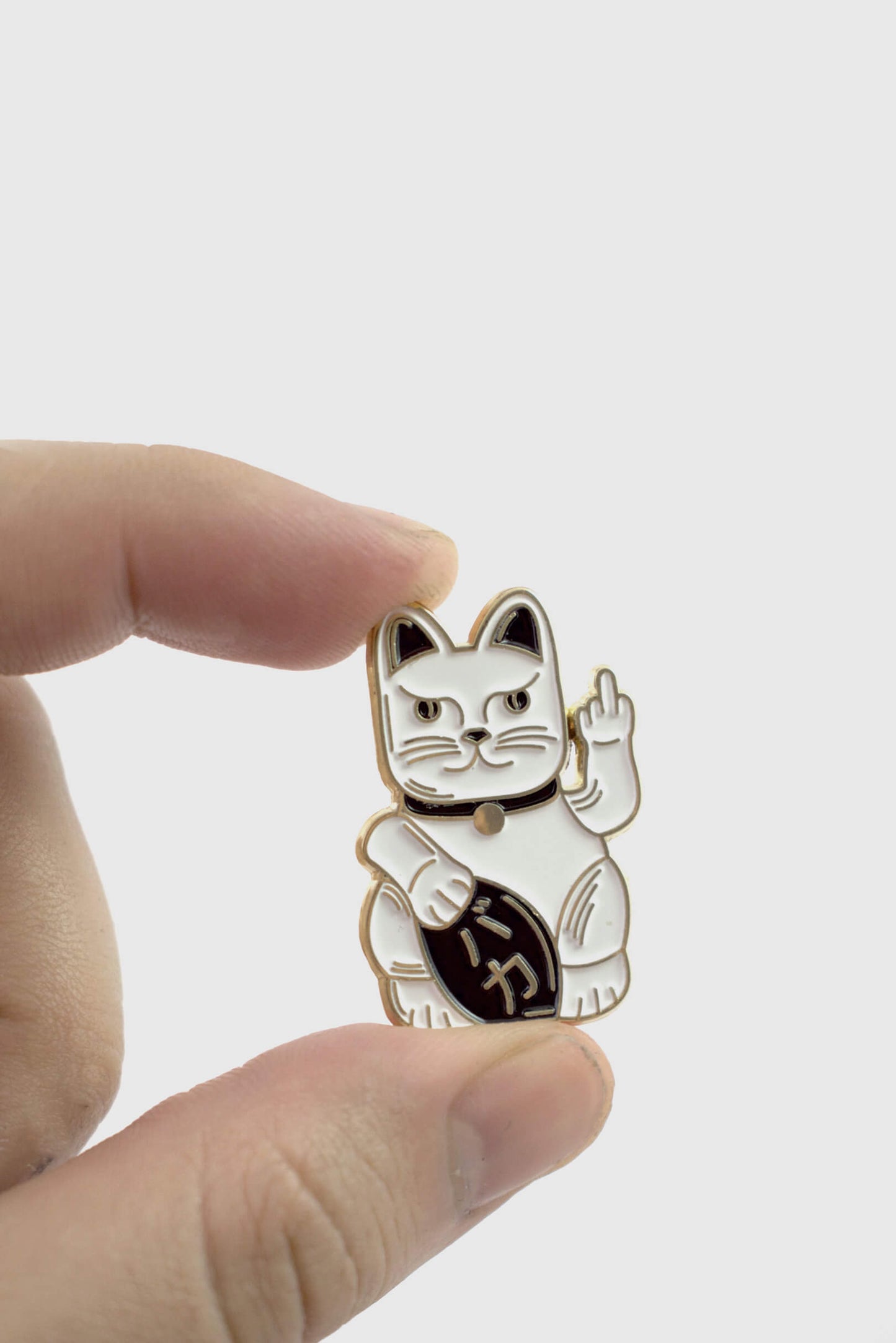 A hand holding the Lucky Cat Pin from Badaboöm Studio, a small enamel pin shaped like a fortune cat whose giving the finger, shown against a white background. 