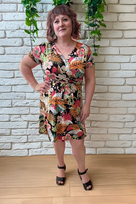 Allana is wearing the Macarena Dress by Rien ne se Perd in Tropical print, a short-sleeved faux-wrap dress with a drawstring waist and a flared above-the-knee skirt. She is standing in front of a white brick wall. 
