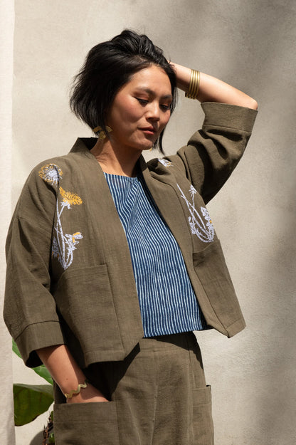 A woman wearing the Magnolia Screen-printed Jacket from Kazak in Olive with Dandelions, a short kimono style jacket with hand-screen printed dandelions, open front, 3/4-length sleeves, and big patch pockets. She is wearing it with matching pants and standing outside by a grey wall. 