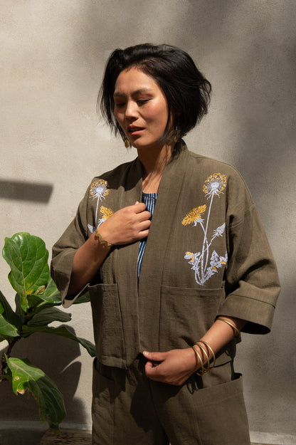 A woman wearing the Magnolia Screen-printed Jacket from Kazak in Olive with Dandelions, a short kimono style jacket with hand-screen printed dandelions, open front, 3/4-length sleeves, and big patch pockets. She is wearing it with matching pants and standing outside by a grey wall with a plant beside her.
