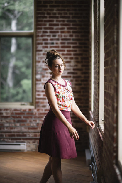 Side view of a woman wearing the Ocean Reversible Dress by Marie C in Cherry. The dress features a patterned bodice with a round neck and a cutout detail, cap sleeves, and a solid coloured skirt and opposite side bodice, with a fit and flare shape. She is standing in a room with a brick wall. 