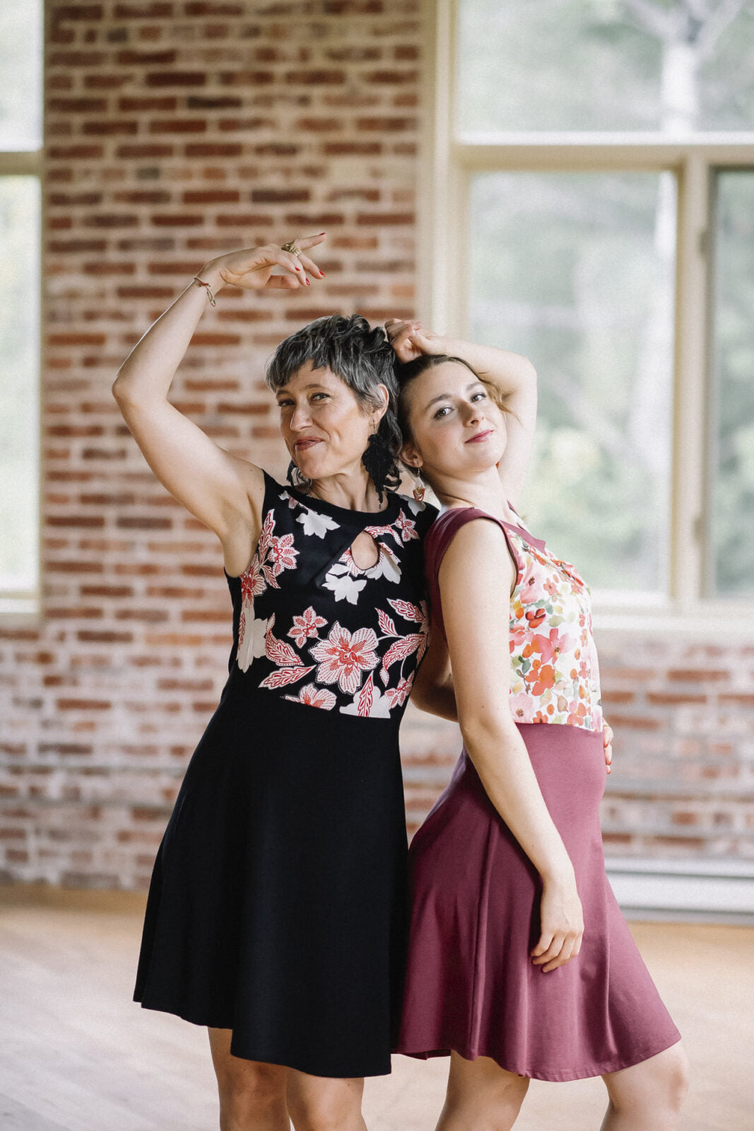 Two women wearing the Ocean Reversible Dress by Marie C, one in Cherry and one in Black. The dress features a patterned bodice with a round neck and a cutout detail, cap sleeves, and a solid coloured skirt and opposite side bodice, with a fit and flare shape. They are standing in a room with a brick wall. 