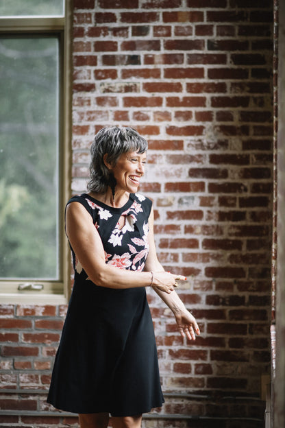 A woman wearing the Ocean Reversible Dress by Marie C in Black. The dress features a patterned bodice with a round neck and a cutout detail, cap sleeves, and a solid coloured skirt and opposite side bodice, with a fit and flare shape. She is standing in a room with a brick wall. 