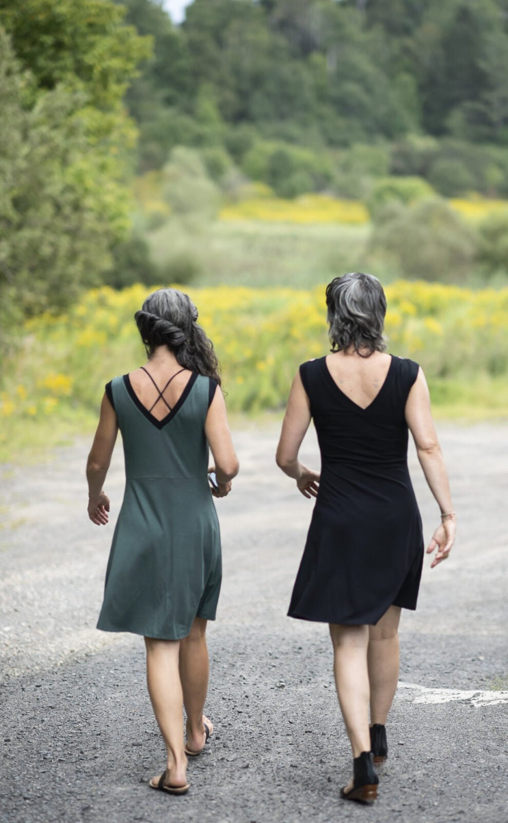 Back view of two women wearing the Ocean Reversible Dress by Marie C. The dress features a patterned bodice with a round neck and a cutout detail, cap sleeves, and a solid coloured skirt and opposite side bodice, with a fit and flare shape. They are standing outside on a gravel road. 