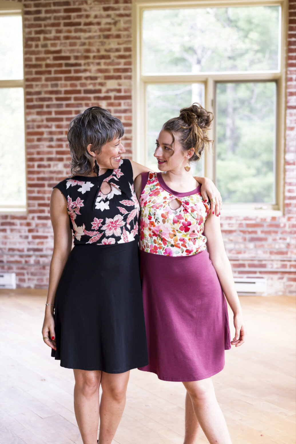 Two women wearing the Ocean Reversible Dress by Marie C, one in Cherry and one in Black. The dress features a patterned bodice with a round neck and a cutout detail, cap sleeves, and a solid coloured skirt and opposite side bodice, with a fit and flare shape. They are standing in a room with a brick wall. 