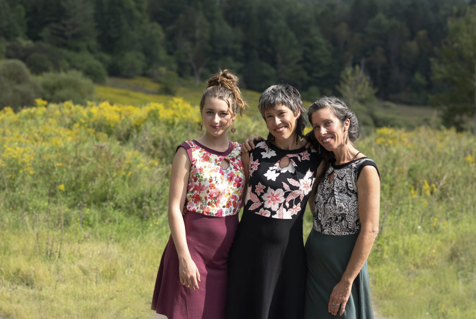 Three women wearing the Ocean Reversible Dress by Marie C. The dress features a patterned bodice with a round neck and a cutout detail, cap sleeves, and a solid coloured skirt and opposite side bodice, with a fit and flare shape. They are standing in a field.
