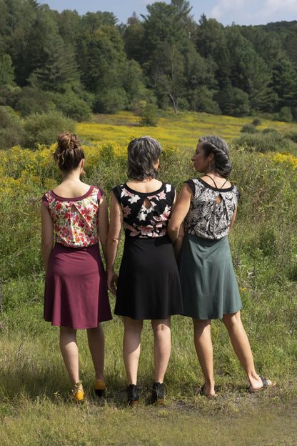 Back view of three women wearing the Ocean Reversible Dress by Marie C. The dress features a patterned bodice with a round neck and a cutout detail, cap sleeves, and a solid coloured skirt and opposite side bodice, with a fit and flare shape. They are standing in a field.
