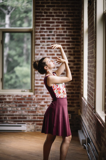 Side view of a woman wearing the Ocean Reversible Dress by Marie C in Cherry. The dress features a patterned bodice with a round neck and a cutout detail, cap sleeves, and a solid coloured skirt and opposite side bodice, with a fit and flare shape. She is standing in a room with a brick wall. 