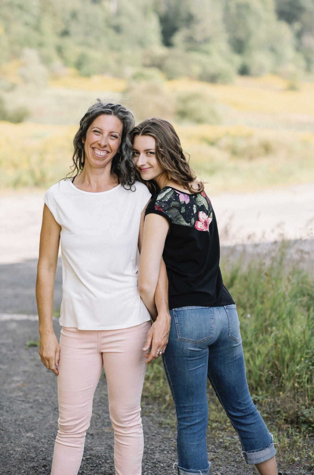 Two women wearing the Marie C Pepita Reversible Top, one in Ivory and one in Black Eyelet. The cap-sleeved top features patterned fabric across the chest on one side with solid colour on the other side. They are standing on a gravel road.
