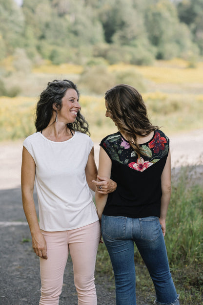 Two women wearing the Marie C Pepita Reversible Top, one in Ivory and one in Black Eyelet. The cap-sleeved top features patterned fabric across the chest on one side with solid colour on the other side. They are standing on a gravel road.
