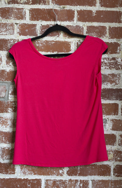 The Sol Top by Marie C in Coral, with a scoop neck and cap sleeves, is shown hanging against a brick wall. 