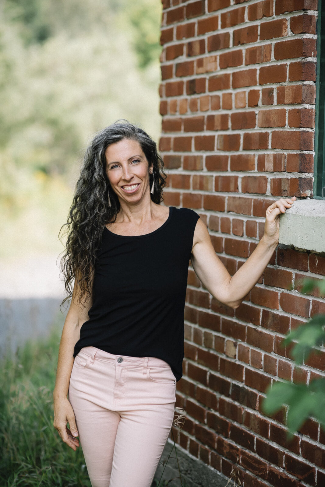 A woman wearing the Sol Top by Marie C in Black, with a scoop neck and cap sleeves, with light pants. She is standing beside a brick house.