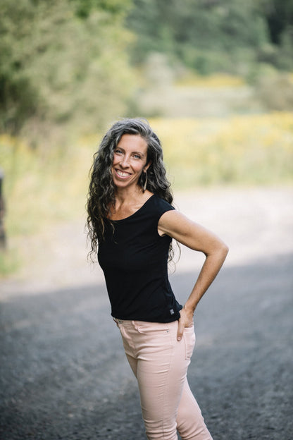 A woman wearing the Sol Top by Marie C in Black, with a scoop neck and cap sleeves, with light pants. She is standing on a gravel road. 