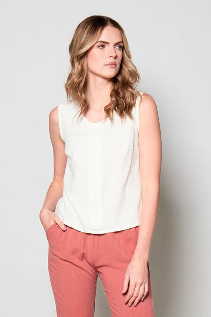 3/4 view of a woman wearing the Marney Tank Top by Tangente in White  with a round neck, full-length centre pleat, and darts at the bust. She is wearing it with coral pants and standing in front of a white background. 