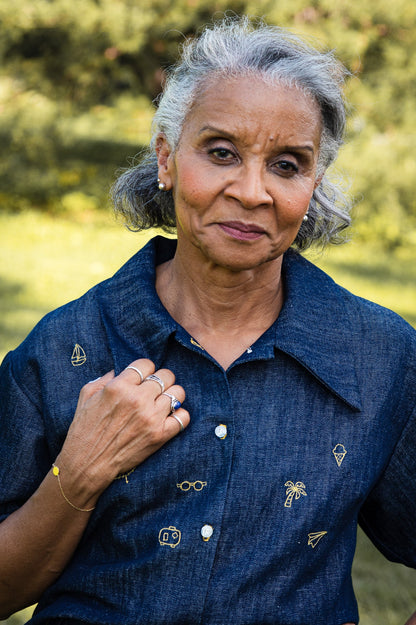 Close-up of a woman wearing the Almeria Blouse by MAS in Denim, a short-sleeved shirt with a large collar, button front, a boxy fit and hand-embroidered designs on the front. She is standing outside. 