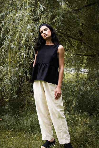 A woman wearing the Malé Tank Top by MAS in Pepper, with a high neck, a curved gathered seam that creates a peplum effect, and three ties at the back. She is wearing it with ivory pants and standing outside. 