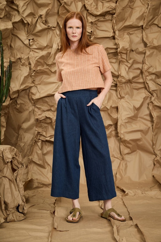 A woman wearing the Mateo Pants by Melow in Denim, ¾ wide leg pants with pull-on waistband that’s elasticized at the back. She is wearing them with an orange top and standing in front of a textured brown background.