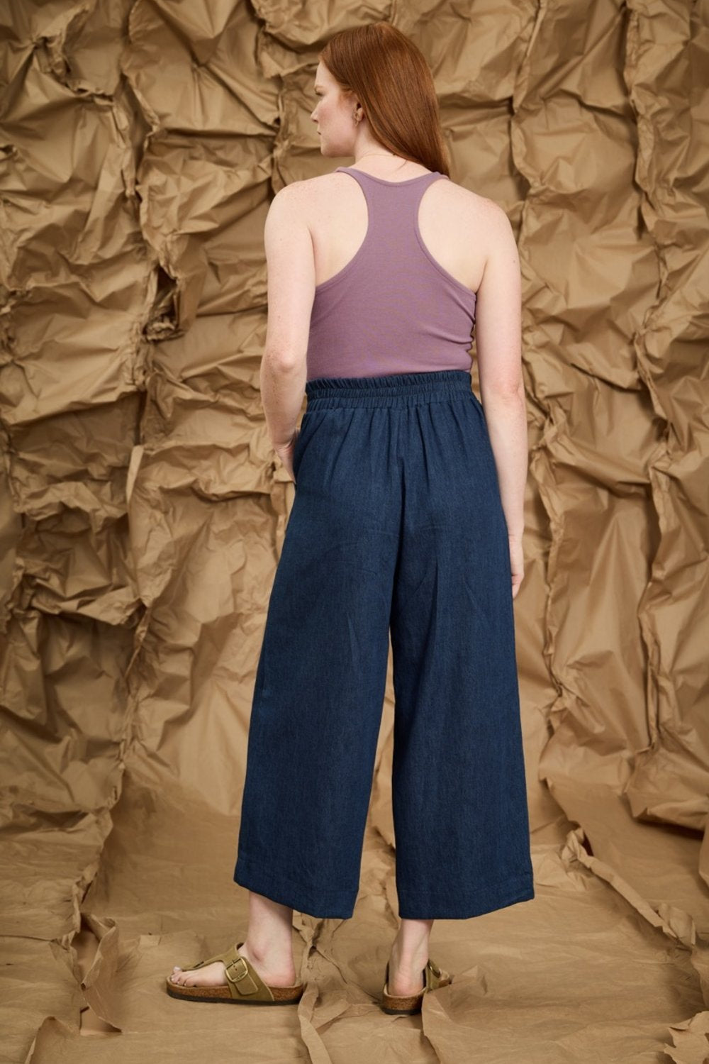 Back view of a woman wearing the Mateo Pants by Melow in Denim, ¾ wide leg pants with pull-on waistband that’s elasticized at the back. She is wearing them with a purple tank top and standing in front of a textured brown background.