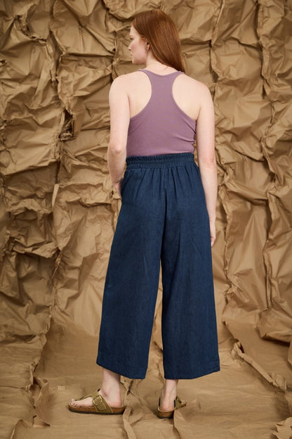 Back view of a woman wearing the Mateo Pants by Melow in Denim, ¾ wide leg pants with pull-on waistband that’s elasticized at the back. She is wearing them with a purple tank top and standing in front of a textured brown background.