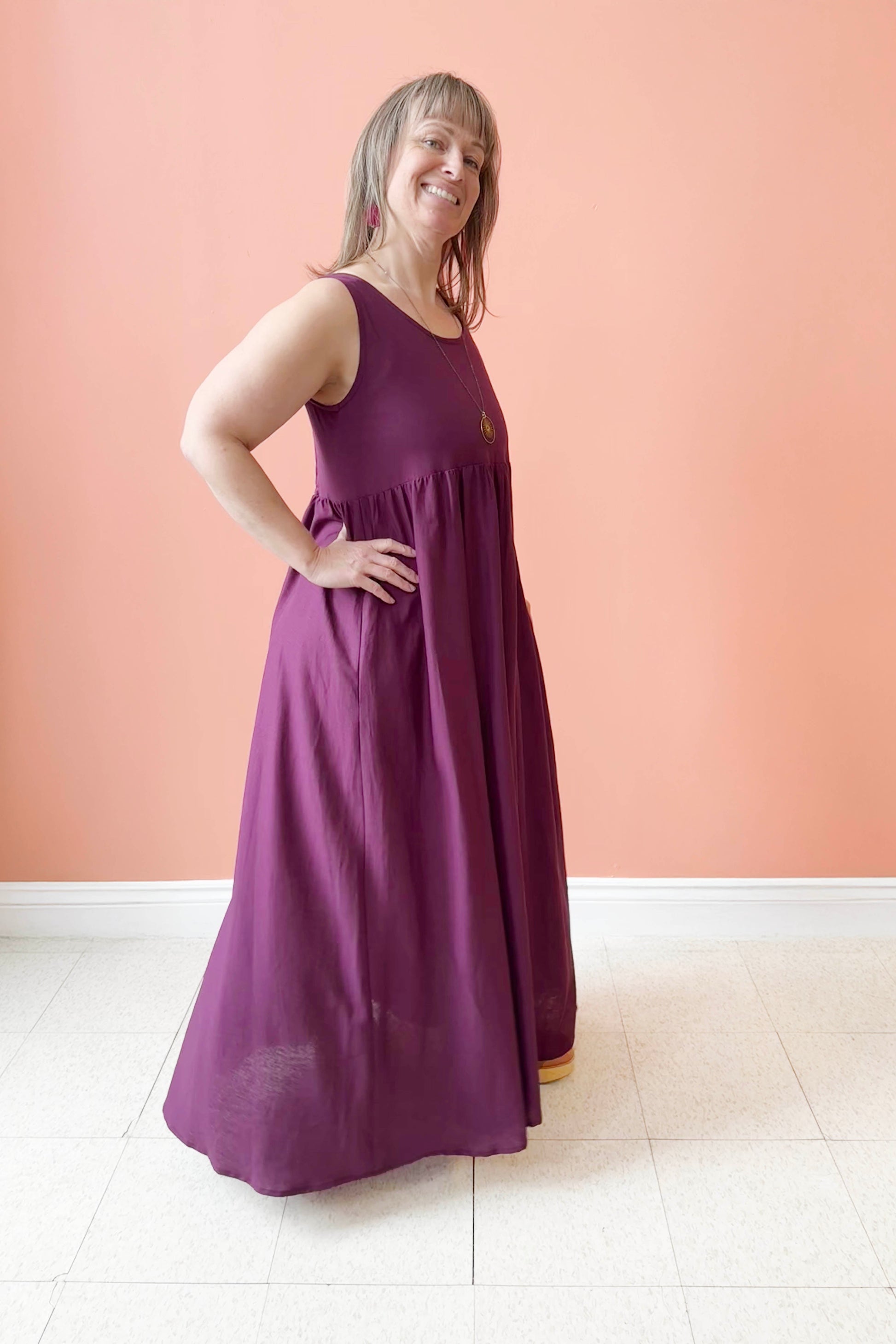 Bridget is wearing the Kim Dress by Melow in Burgundy, a voluminous maxi dress with wide tank straps, a round neck, an empire waist, an honeycomb elastic smocking at the back. She is standing in front of a coral background. 