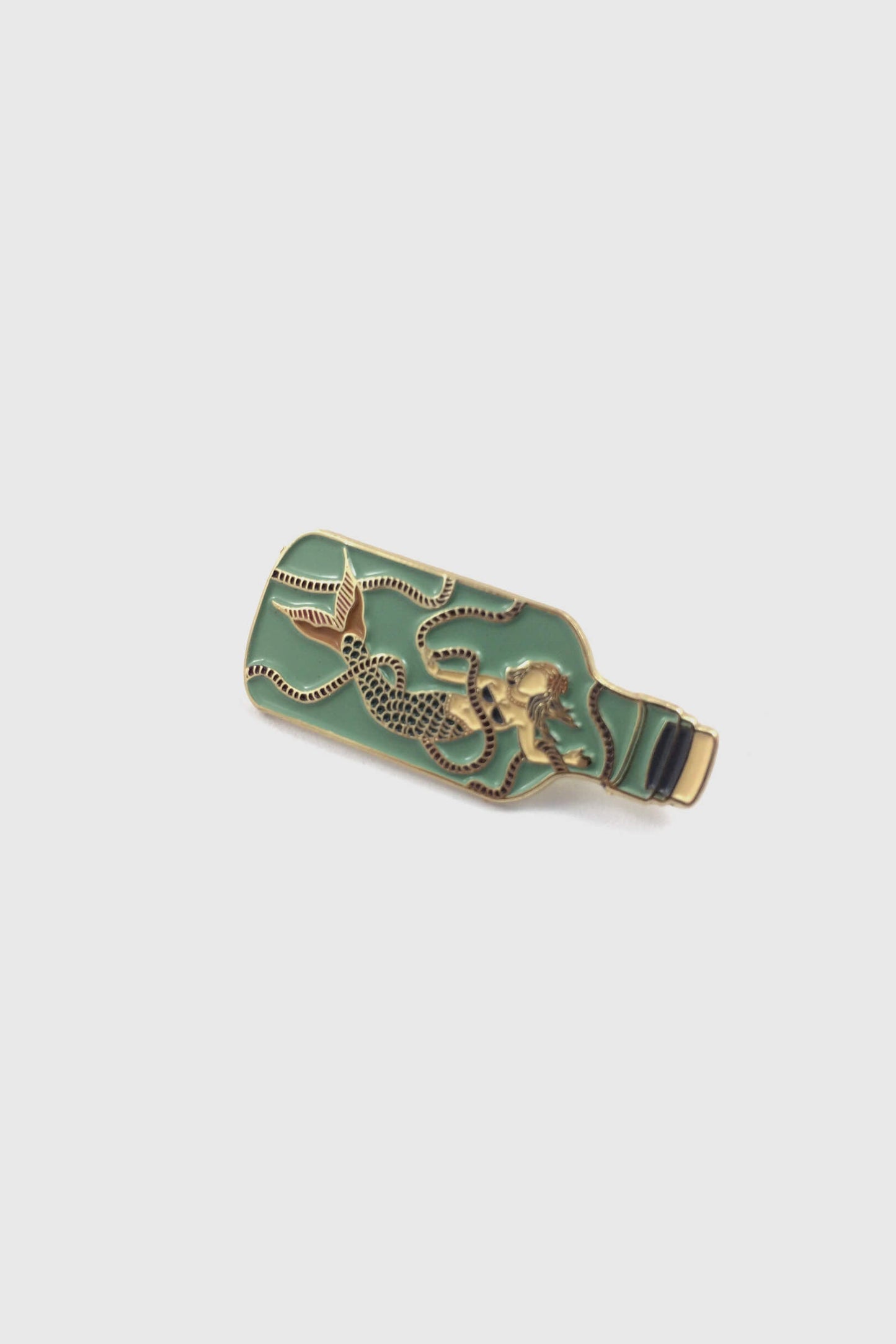 The Mermaid in a Bottle Pin from Badaboöm Studio, an enamel pin featuring a mermaid trapped in a light green bottle, is shown against a white background. 