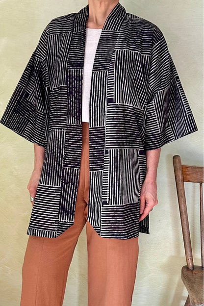 Neck down view of a woman wearing the Mirano Jacket by Kazak in Oris Slate, a cross hatch pattern. It is a  mid-thigh length open kimono jacket with 3/4 sleeves. She is wearing it with the Oxalis pants in Ginger and standing in a room by a wooden chair. 