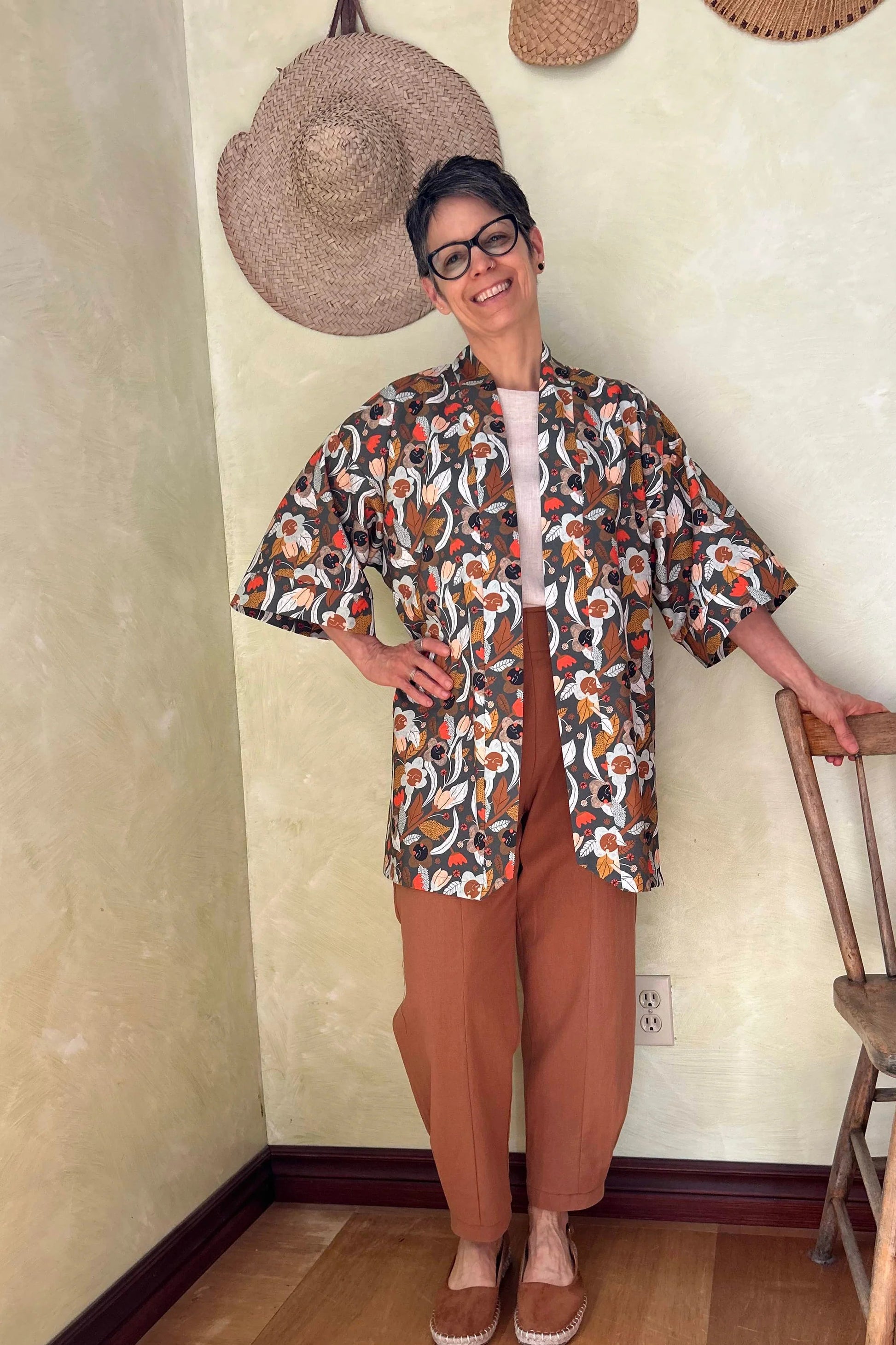 A woman wearing the Mirano Jacket by Kazak in Olive Floral, a mid-thigh length open kimono jacket with 3/4 sleeves. She is wearing it with the Oxalis pants in Ginger and standing in a room by a wooden chair. 