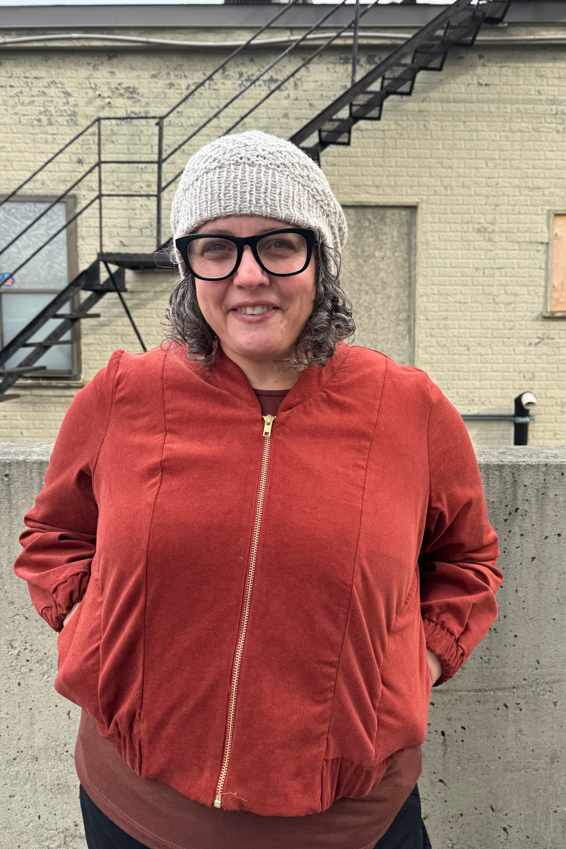 A woman wearing the Beret by Charlevoix Pure Laine in 
Grey with the Redrawn Zip Jacket by Eve Lavoie in Terracotta Corduroy. She is standing outside. 