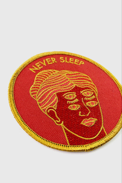 The Never Sleep Patch from Badaboöm Studio, circular embroidered patch with a red background, gold border, and a stylized face with two sets of open eyes. The words 'NEVER SLEEP' are embroidered in gold at the top.