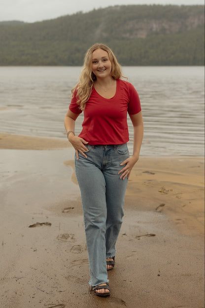 A woman wearing the Rideau Shoreline tee by Blondie in Poppy, with a scoop neck and short sleeves. She is wearing it with jeans and standing on a beach.