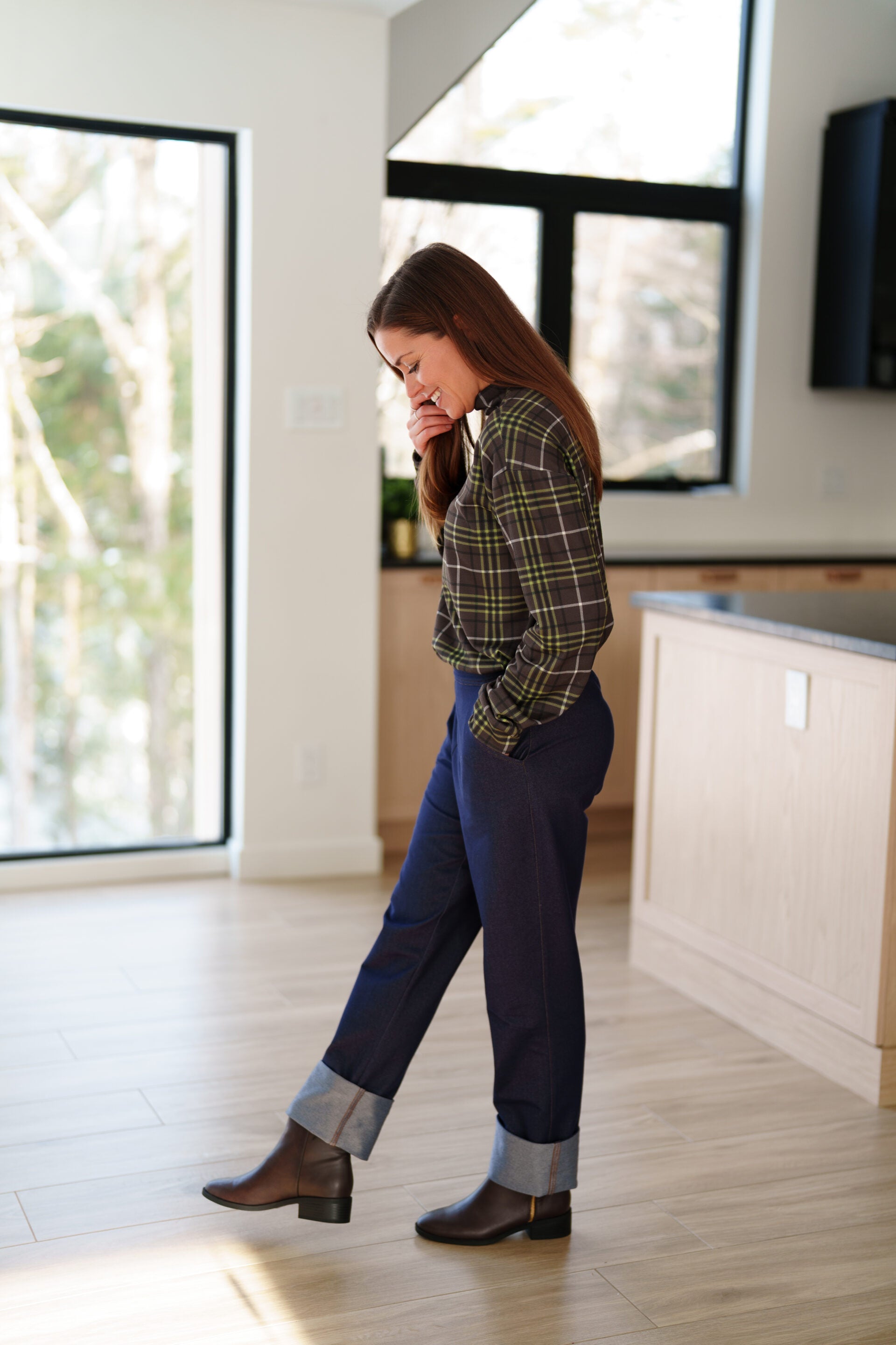 Side view of a woman wearing the Yamaska Pants by Rien ne se Perd in Denim Blue, pull on style jeans with rolled cuffs. She is wearing them with a green and brown plaid shirt, and dark brown ankle boots in a bright room with light wood flooring