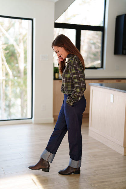 Side view of a woman wearing the Yamaska Pants by Rien ne se Perd in Denim Blue, pull on style jeans with rolled cuffs. She is wearing them with a green and brown plaid shirt, and dark brown ankle boots in a bright room with light wood flooring