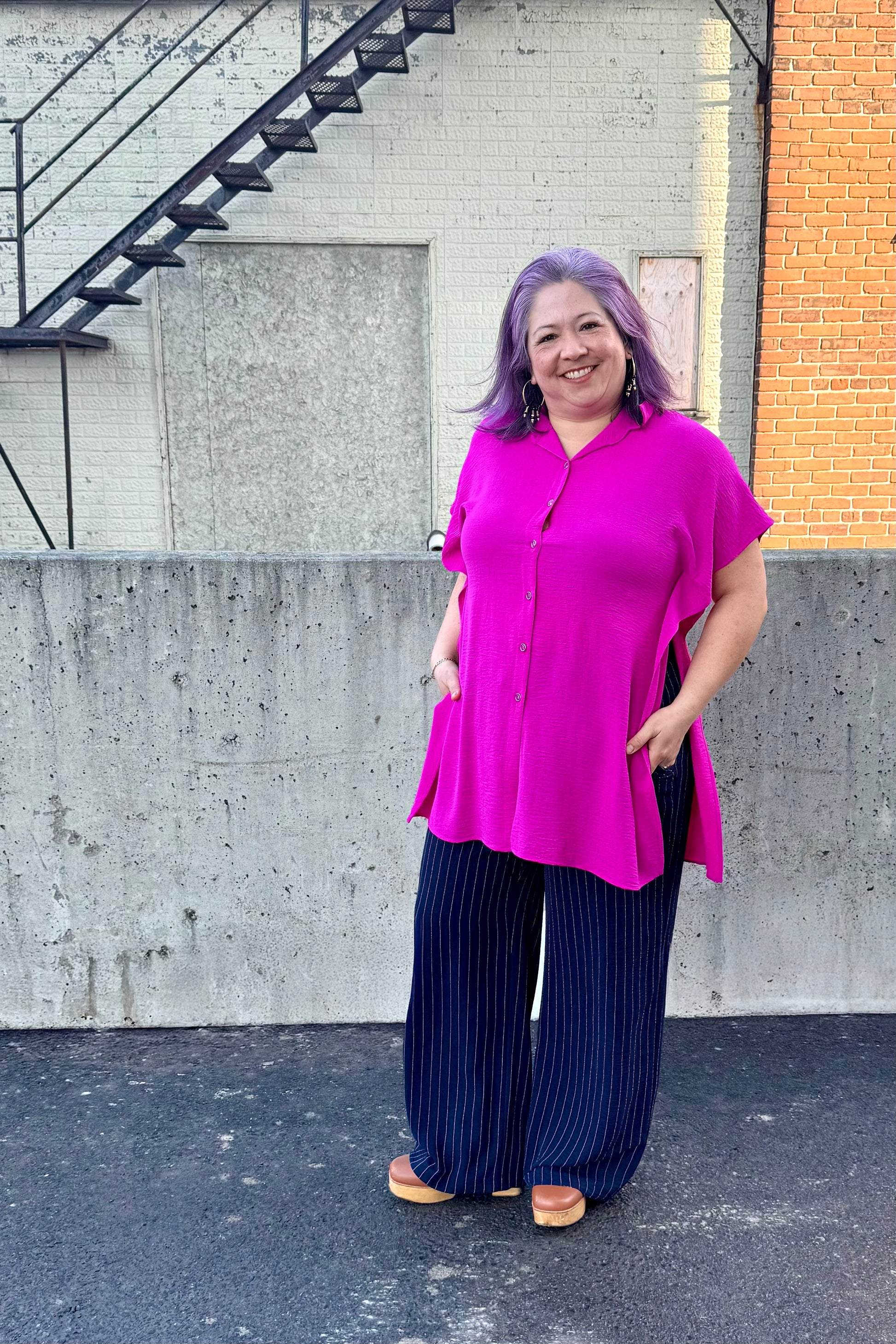 A woman wearing the Geneva Top by Pure Essence in Magenta, a long and loose button front shirt with short sleeves, a classic collar, and high side slits. She is wearing it with the Gabby Pants in navy/white pinstripe and standing outdoors against a concrete wall. 