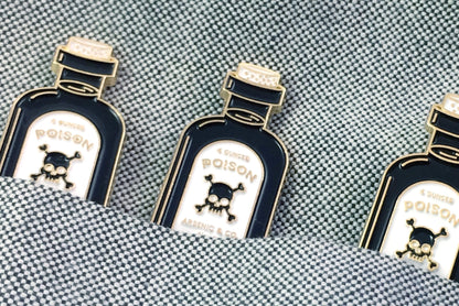 Three Poison Pins by Badaboöm Studio, featuring a tiny bottle of poison, are shown tucked into a textured blue pocket. 