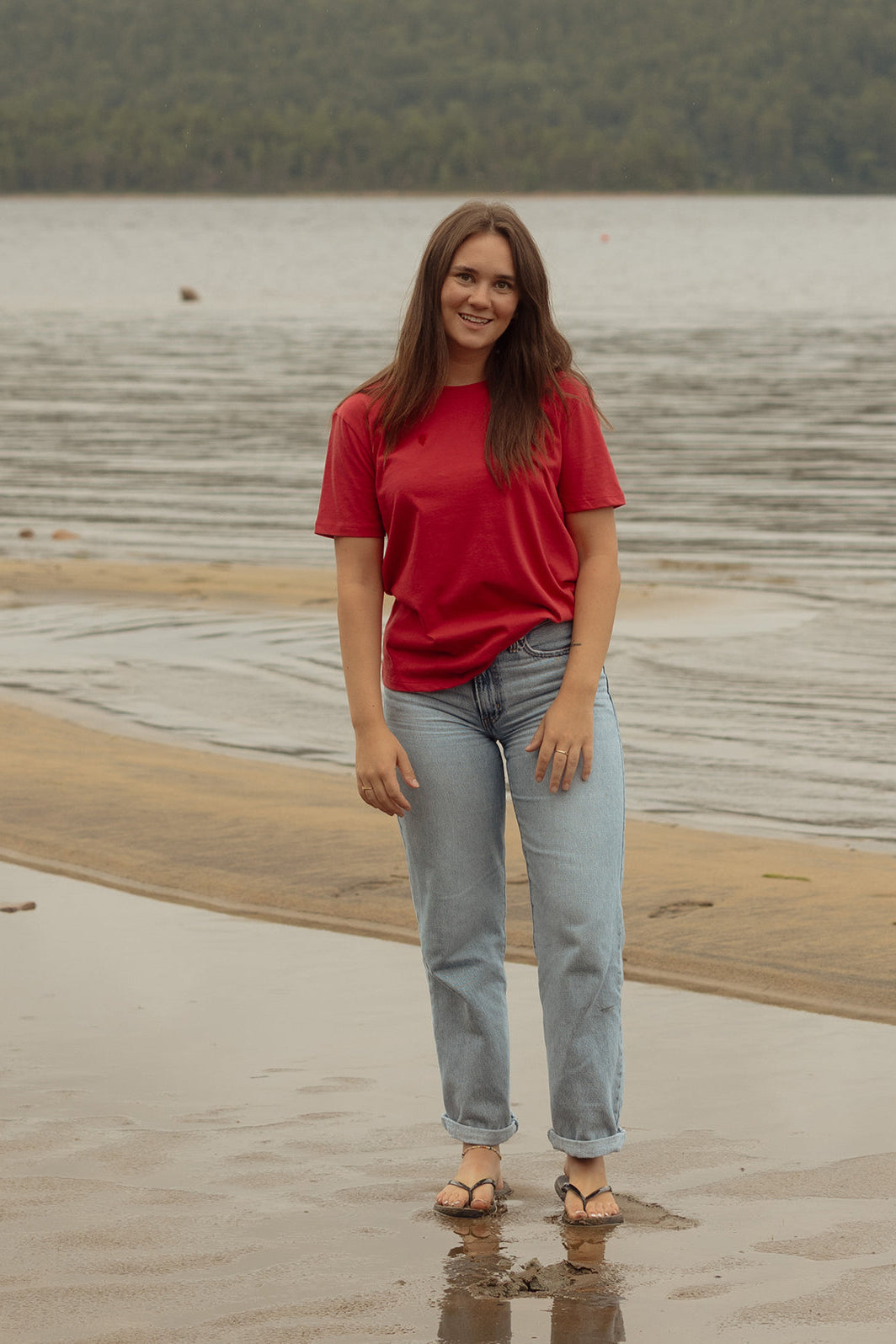 A woman wearing the Prairie Tee by Blondie in Poppy with jeans, standing on a beach 