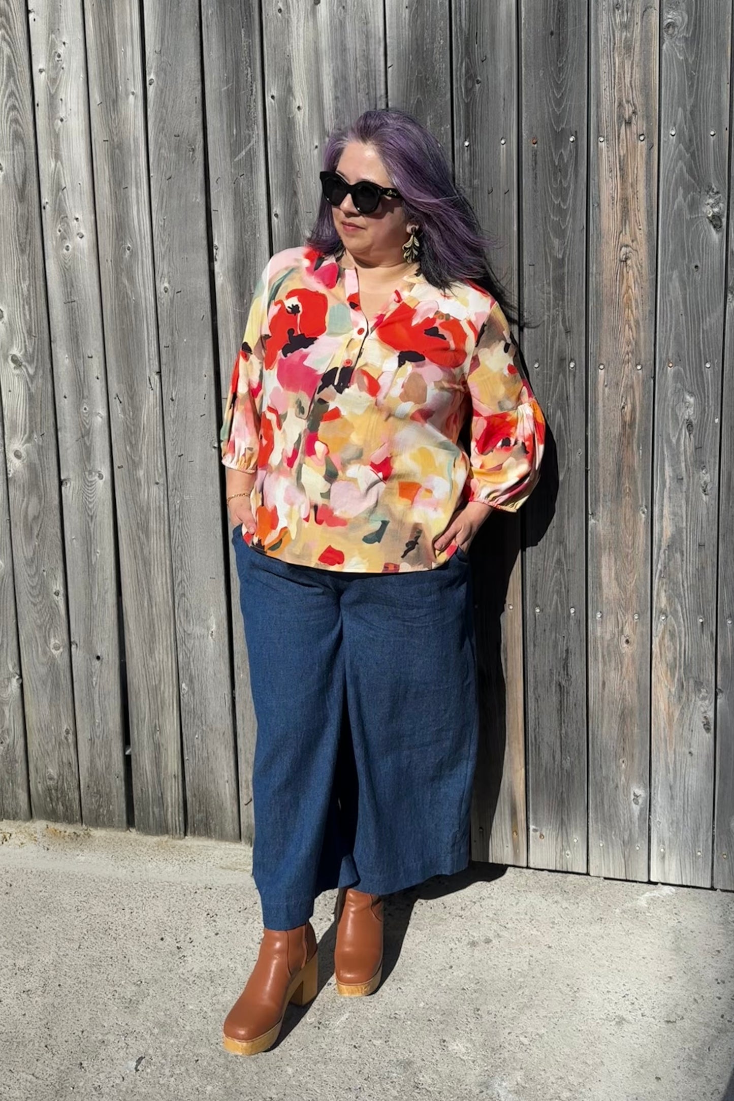 A woman wearing the Mateo Pants by Melow in Denim, ¾ wide leg pants with pull-on waistband that’s elasticized at the back. She is wearing them with a floral top and leaning against a wooden fence. 