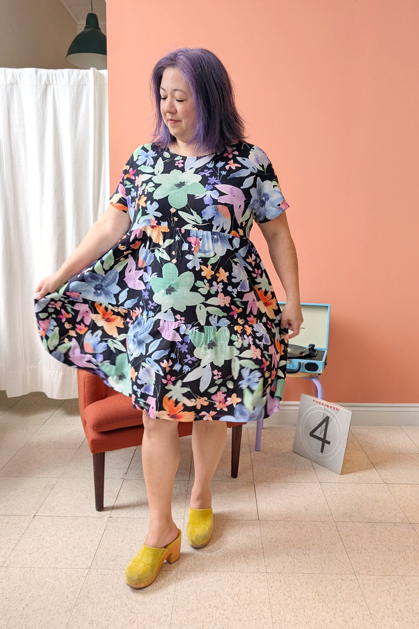 Woman wearing the Grayson Dress by Pure Essence in Moonlit Bloom, a knee-length baby doll style dress with a tiered skirt and short sleeves. She is standing in a room with a peach wall and white curtain