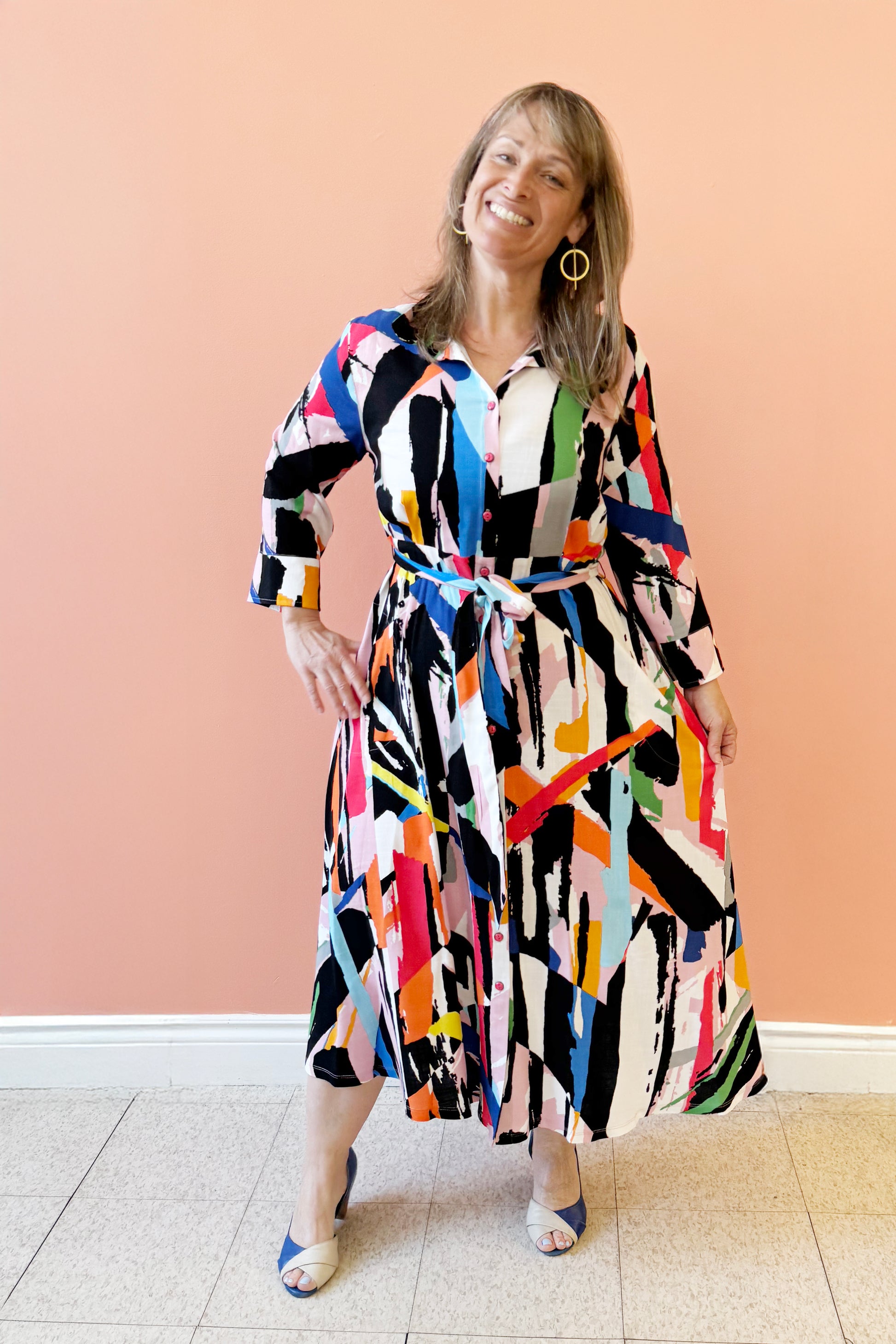 Bridget is wearing the Elliott Dress by Pure Essence in Multicolour geometric print, a long sleeved shirt dress with a button front, blouse collar, tie-belt at the waist, and full maxi skirt. She is standing in front of a coral wall. 