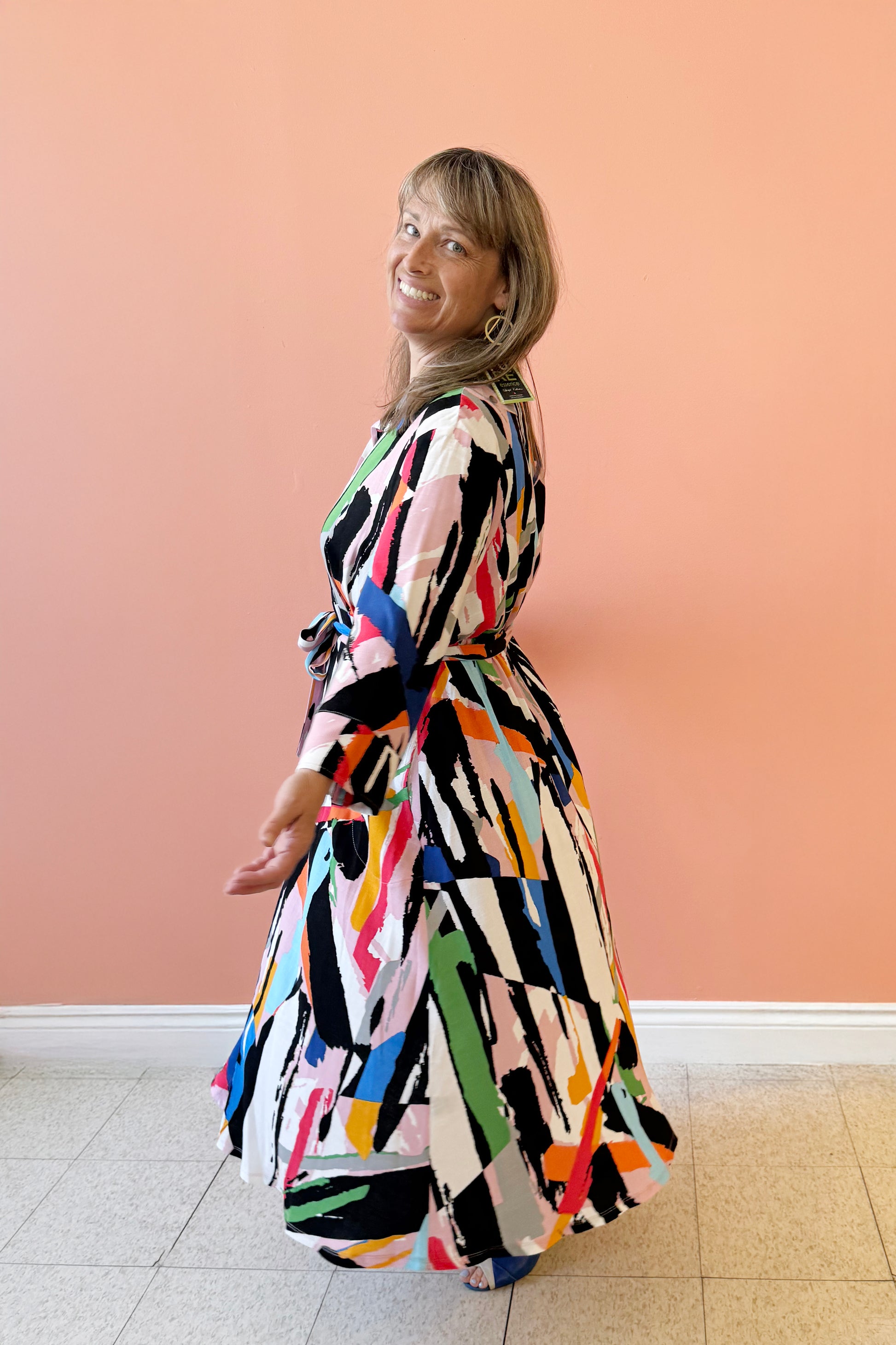Side view of Bridget wearing the Elliott Dress by Pure Essence in Multicolour geometric print, a long sleeved shirt dress with a button front, blouse collar, tie-belt at the waist, and full maxi skirt. She is standing in front of a coral wall. 