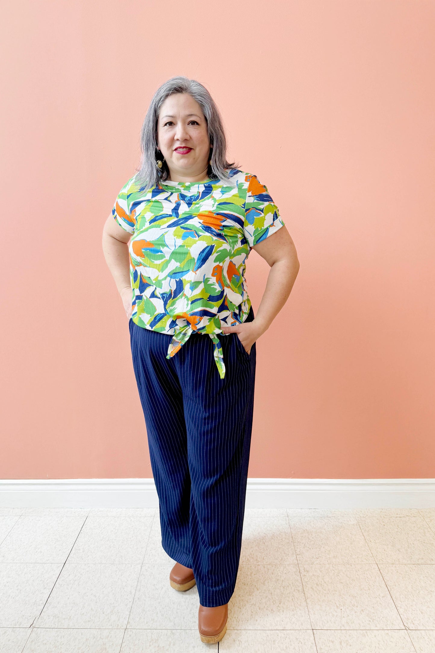 Christina is wearing the Ellen Tunic by Pure Essence in Blue/Cilantro, a short-sleeved top with a round neck and a tie detail at the waist. She is wearing it with the Eloise Pants in Ink pinstripe and is standing in front of a coral background. 