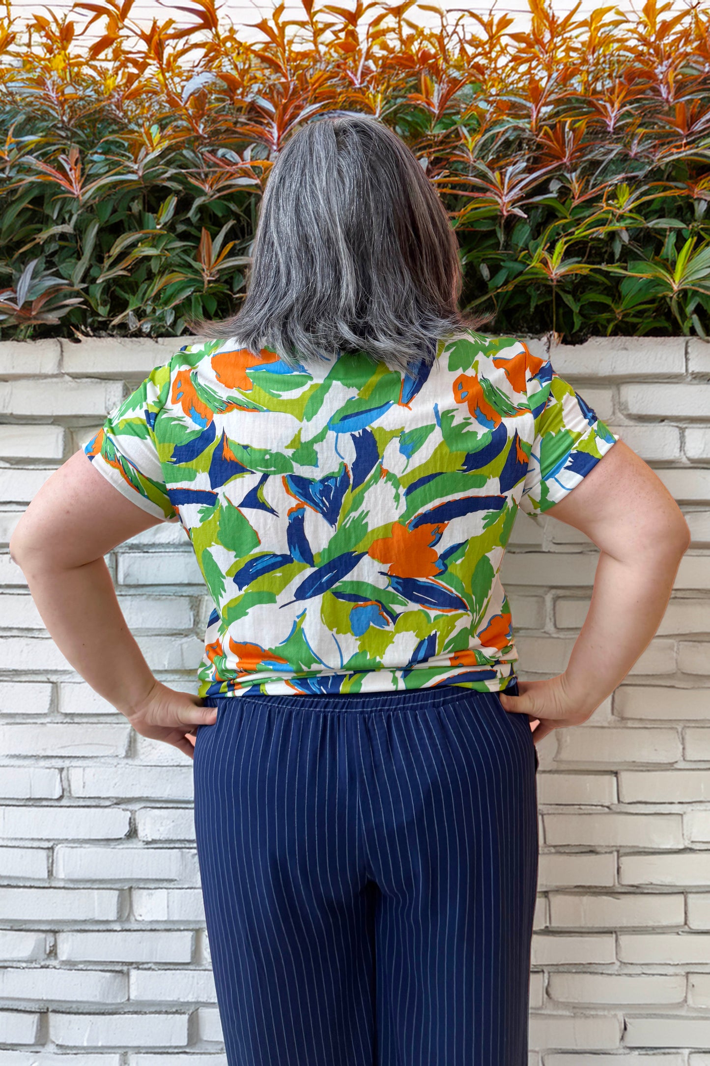 Back view of Christina wearing the Ellen Tunic by Pure Essence in Blue/Cilantro, a short-sleeved top with a round neck and a tie detail at the waist. She is wearing it with the Eloise Pants in Ink pinstripe and is standing in front of white brick wall. 