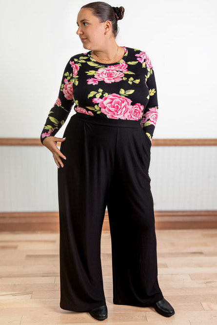 A woman wearing the Cleo Pants by Slak in Black, wide legged pants with subtle pleats and a smooth elastic waist. She is wearing them with a floral sweater and standing in a white room with a wooden floor and wooden wainscotting.