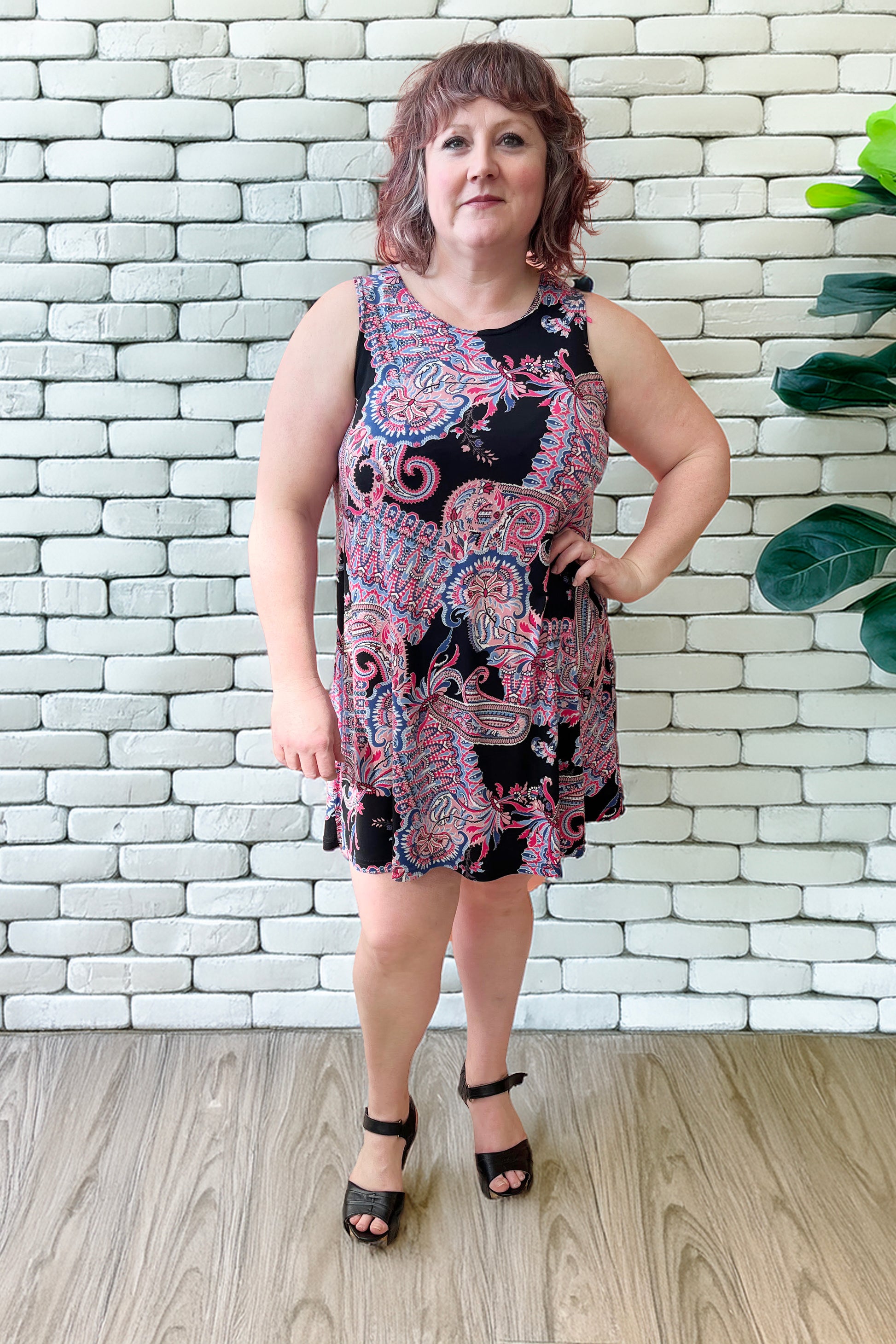 Allana is wearing the Musette Dress by Rien ne se Perd, a short sleeveless swing dress with a purple Paisley print. She is standing in front of a white brick wall. 