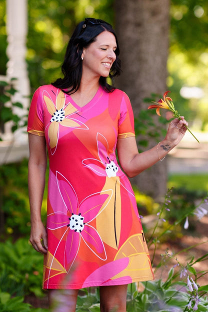 A woman wearing the Aperol Dress from Rien ne se Perd, a short flared dress with a V-neck, short sleeves, and a colourful Pink floral pattern She is holding a flower in a garden setting.